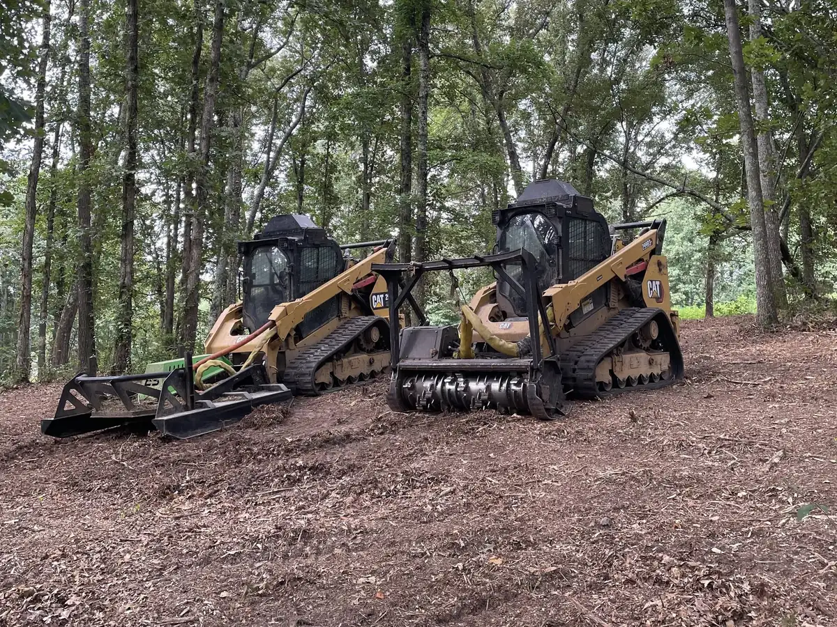 Two tracked machines on a job site, one carrying a forestry mulching head and one a grapple rake, on ground already mulched
