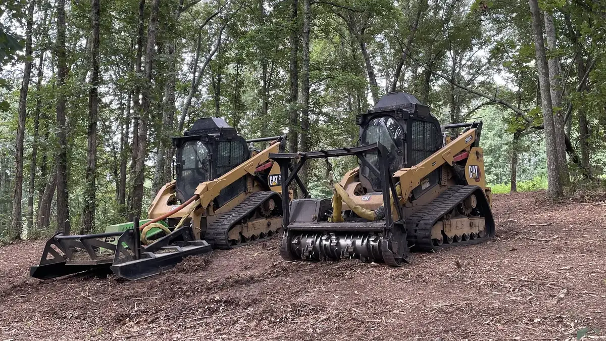 A pair of tracked skid steers parked on freshly mulched ground under standing timber